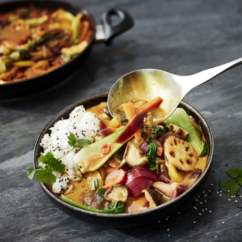 Gemüsecurry mit Reis, Kräutern und buntem Gemüse in einer Fissler-Schüssel; ein Präzisionslöffel hebt eine Portion an. Langlebiges Kochgeschirr sichtbar.