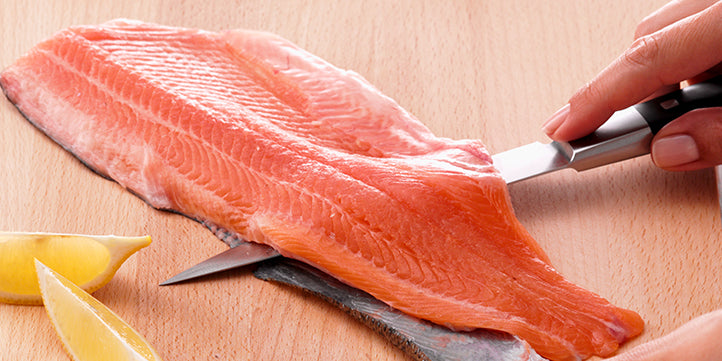 A person is filleting fresh salmon on a wooden board with lemon slices, using a Fissler premium knife for precision and quality.