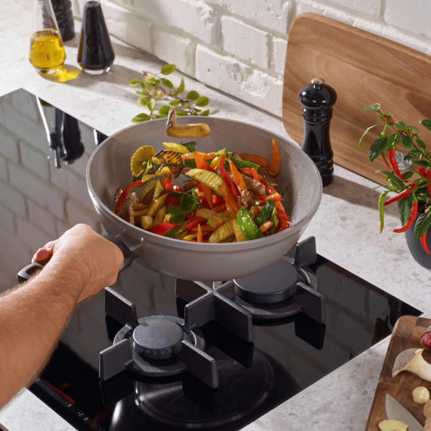 Eine Hand kocht lebhaftes Pfannengemüse in einer Fissler-Edelstahlpfanne auf einem Gasherd; hochwertige Zutaten auf dem Tresen.