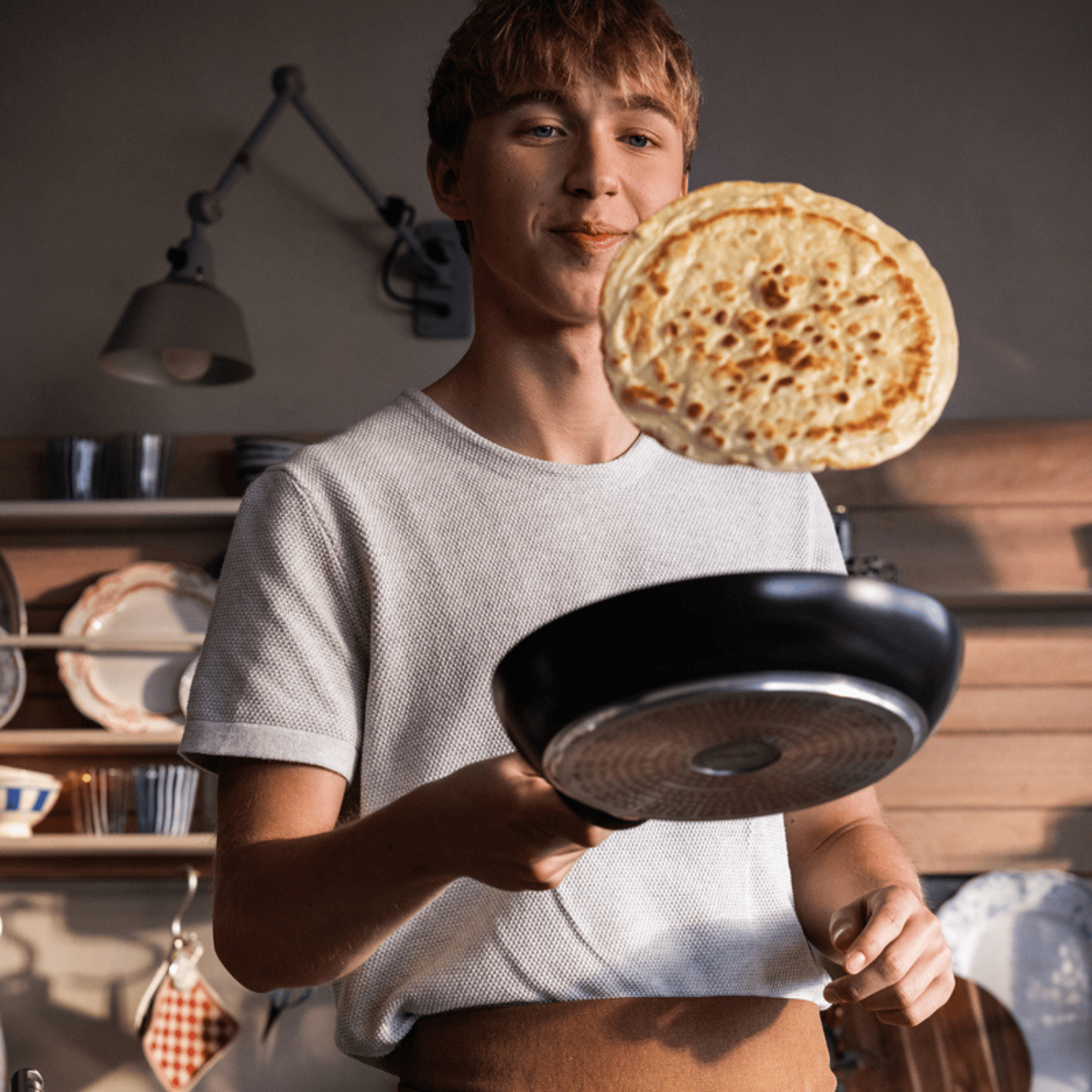 Eine junge Person wendet einen Pfannkuchen in einer hochwertigen Fissler-Pfanne und präsentiert deutsche Qualität in einer gemütlichen Küche mit Regalen und Tellern.