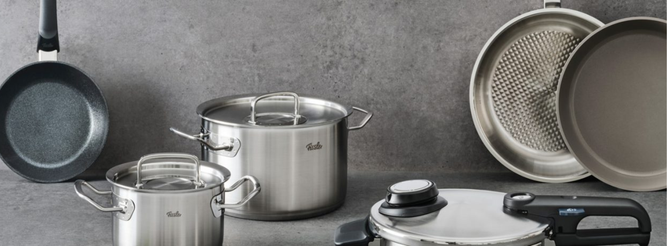 Fissler Edelstahl-Kochgeschirr-Set: Töpfe, Pfannen und Schnellkochtopf auf grauer Arbeitsplatte, die deutsche Präzision verkörpern.