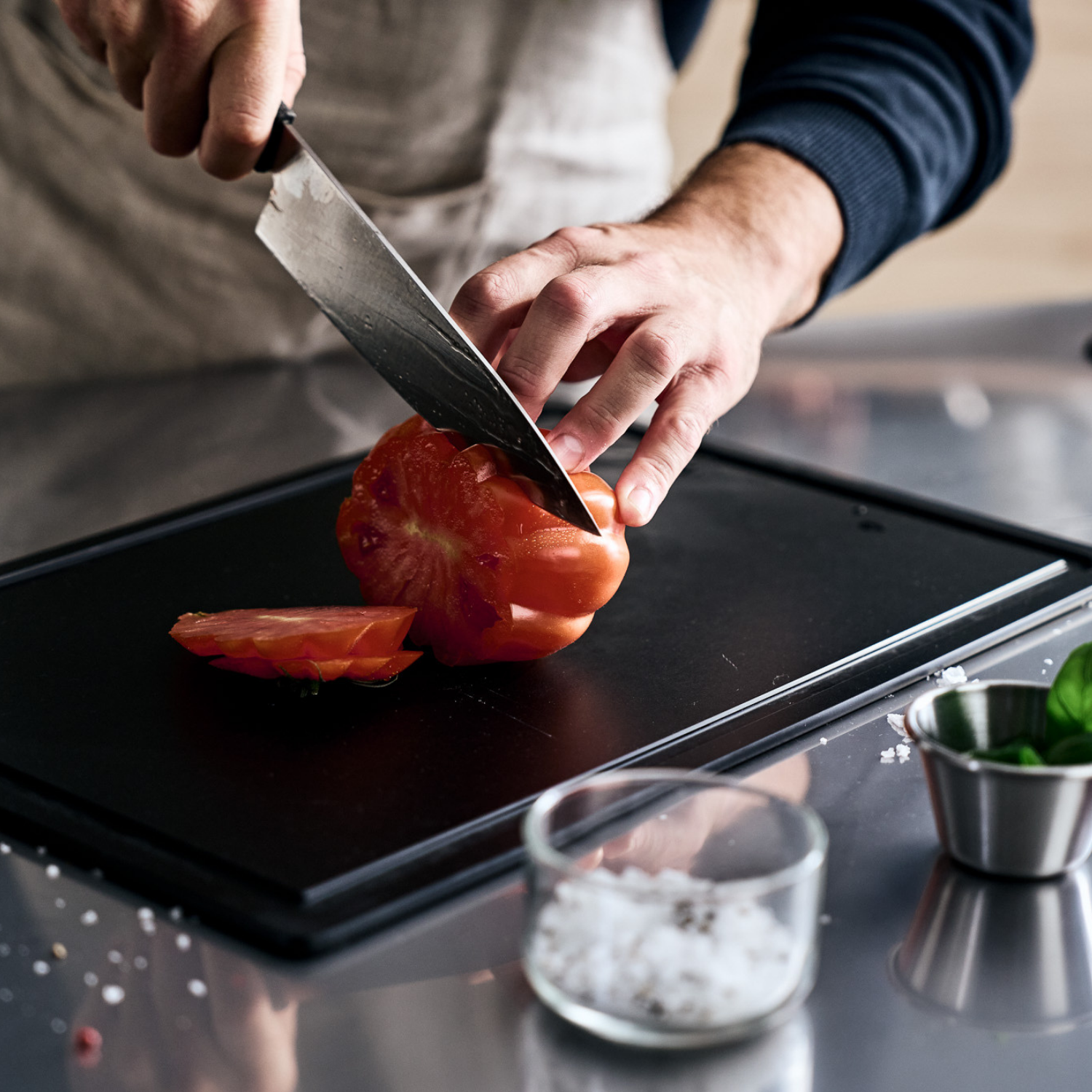 A chef cuts a tomato on an elegant Fissler cutting board; coarse salt, basil, and scattered seeds lie on a polished stainless steel counter.