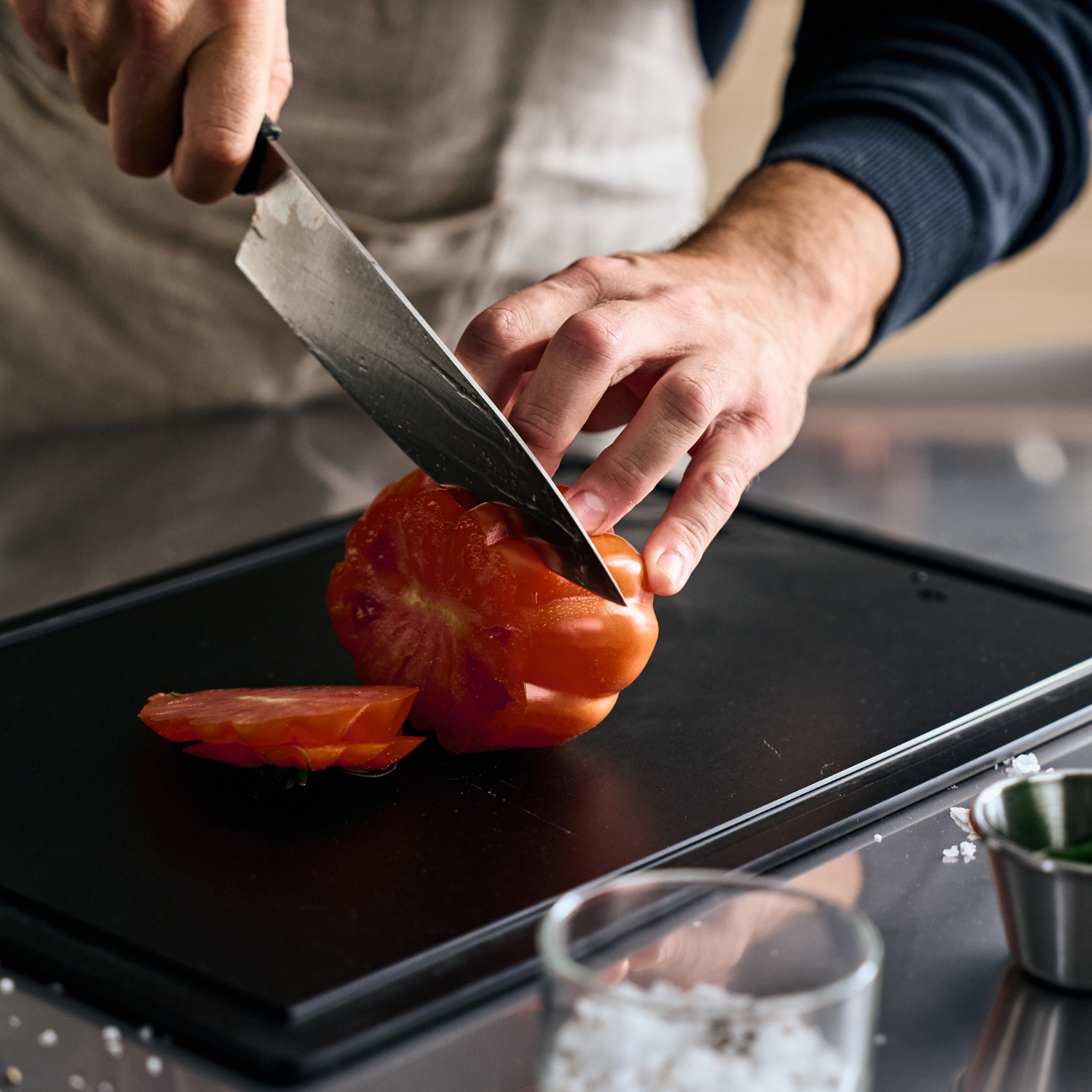 Eine Person in einer Schürze schneidet eine Tomate auf einem Fissler-Schneidebrett präzise in Scheiben, während Salz und Kräuter in stilvollen Schälchen zur Hand sind.