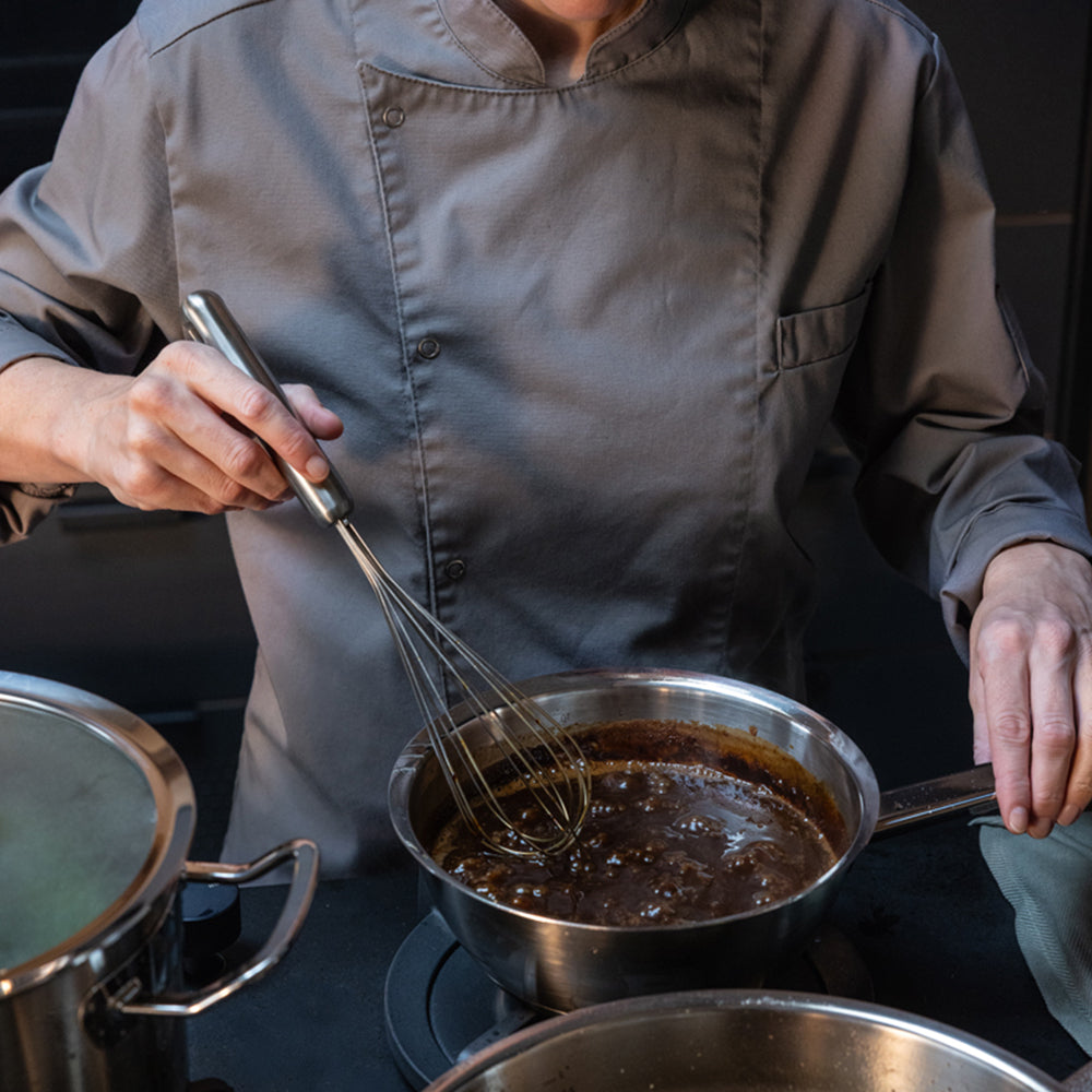 A chef in a grey uniform expertly stirs a rich sauce with a whisk in a premium Fissler stainless steel pot on the stovetop, showcasing precision craftsmanship and timeless German design. Another Fissler pot sits nearby, reflecting the brand’s commitment to innovation and durability in professional kitchens.