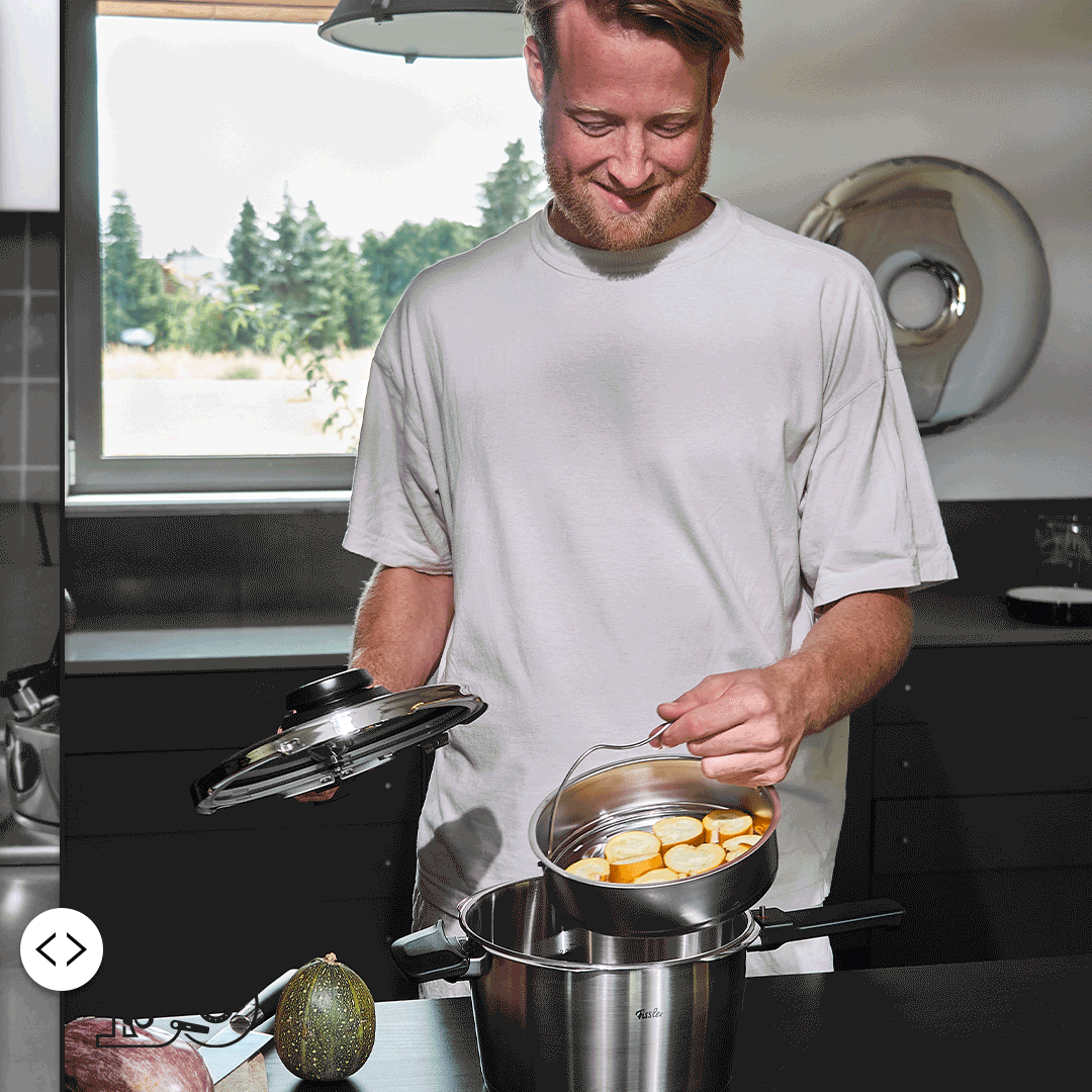 Ein Mann lächelt, während er einen Dampfgarer mit Gemüse in einen Fissler-Edelstahltopf auf eine moderne Küchenzeile stellt. Draußen grünt es.