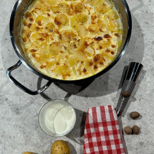 Ein Fissler-Edelstahltopf mit goldenem Kartoffelgratin auf einer grauen Fläche, daneben Küchengeräte und frische Zutaten.