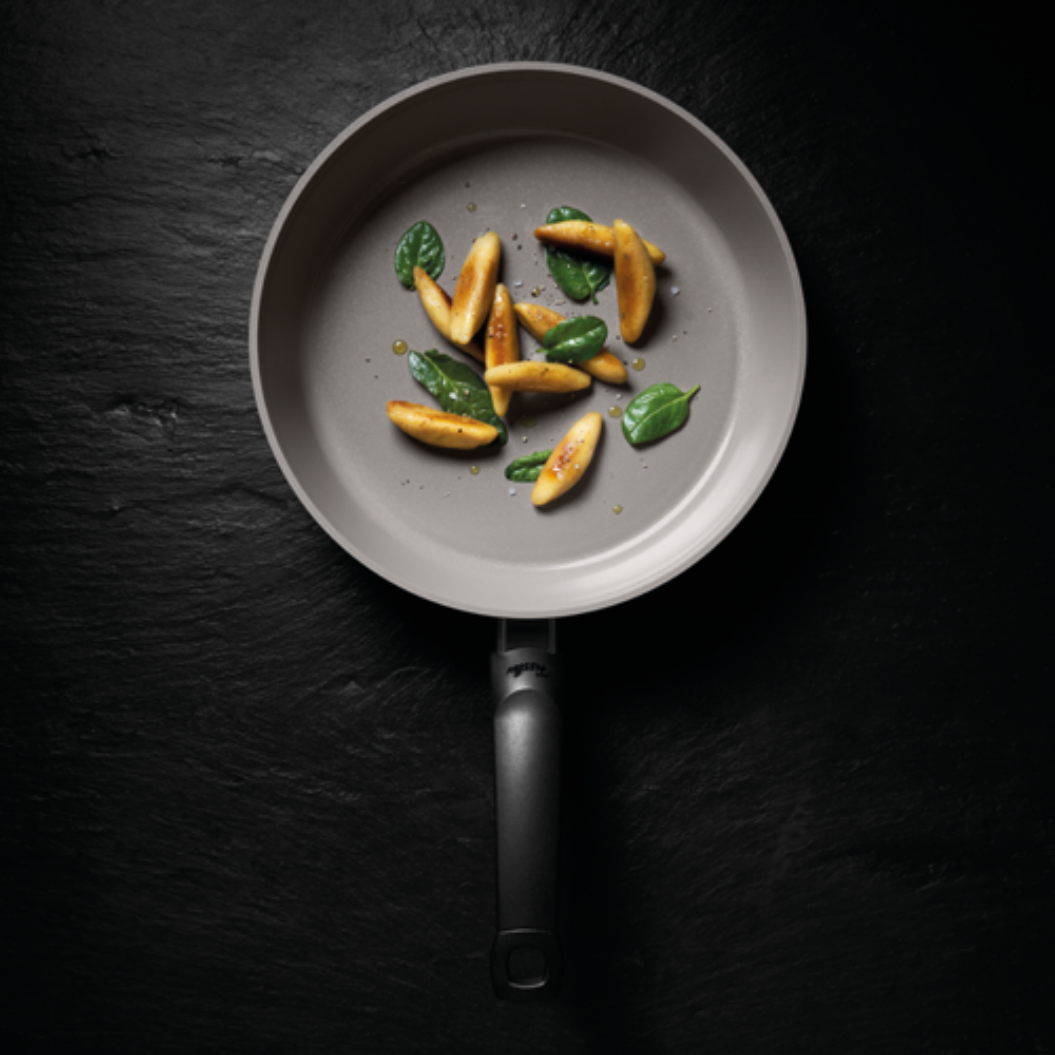 Fissler Antihaft-Pfanne auf dunkler Oberfläche mit goldenen Gnocchi und frischem Basilikum, die deutsche Präzisionsarbeit zeigt.