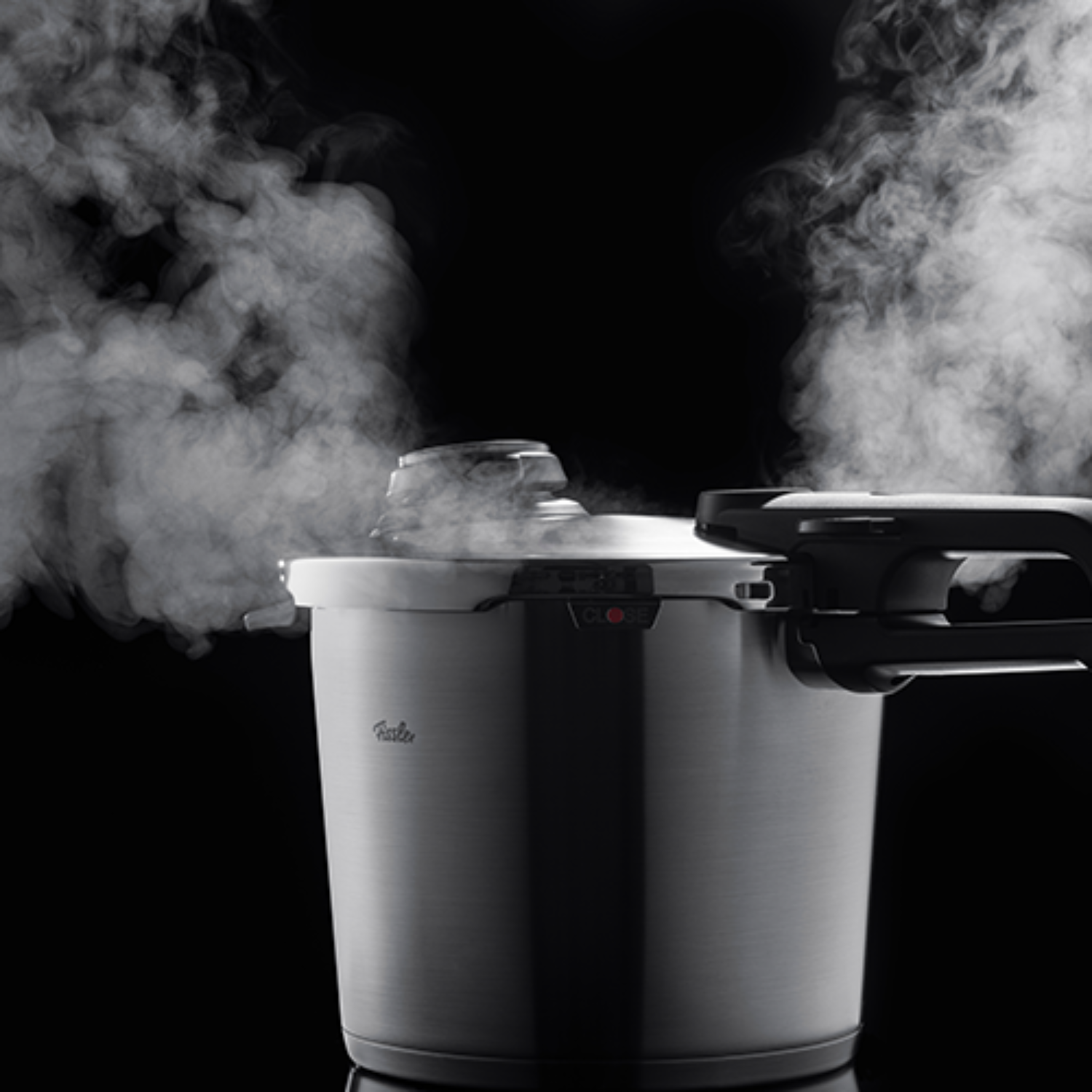 Ein Schnellkochtopf von Fissler aus Edelstahl lässt Dampf ab und präsentiert deutsche Technik und innovatives Design auf schwarzem Hintergrund.