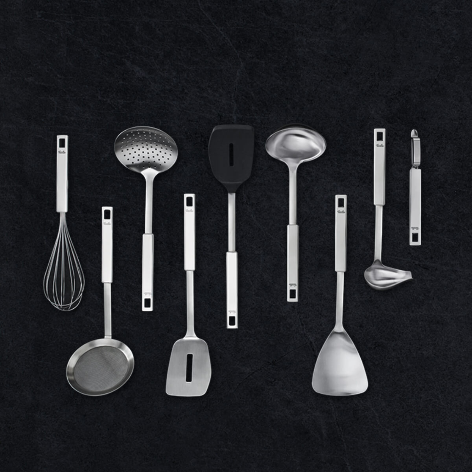 Neun Fissler Küchenwerkzeuge aus rostfreiem Stahl, präzisionsgefertigt in Deutschland, elegant angeordnet auf einer dunkel strukturierten Oberfläche.