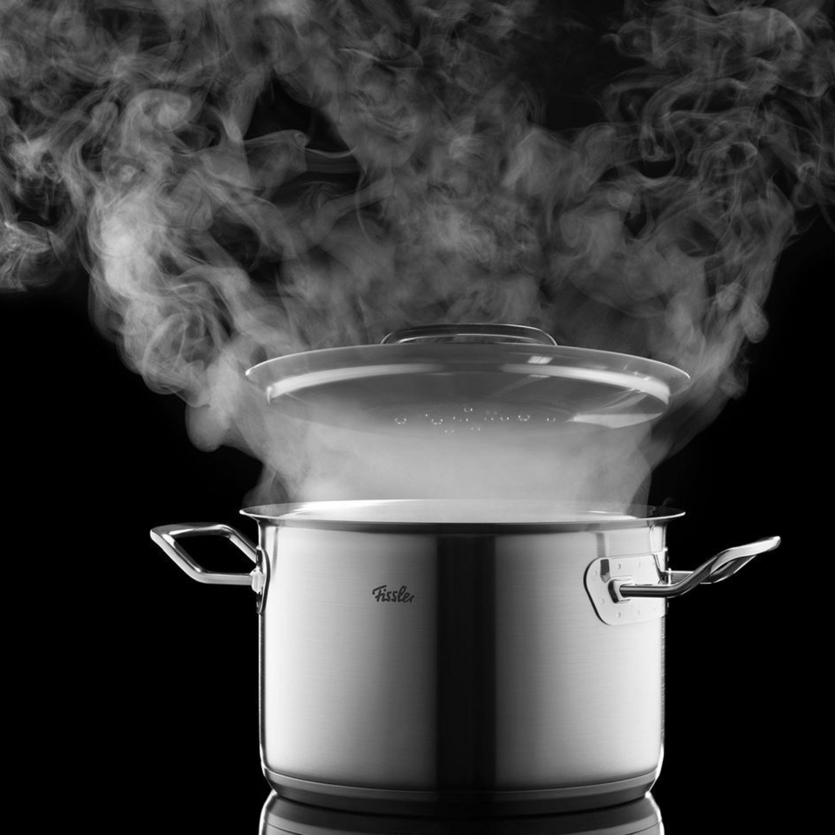 Ein in Deutschland hergestellter Fissler-Edelstahltopf mit Glasdeckel setzt eleganten Dampf vor schwarzem Hintergrund frei.