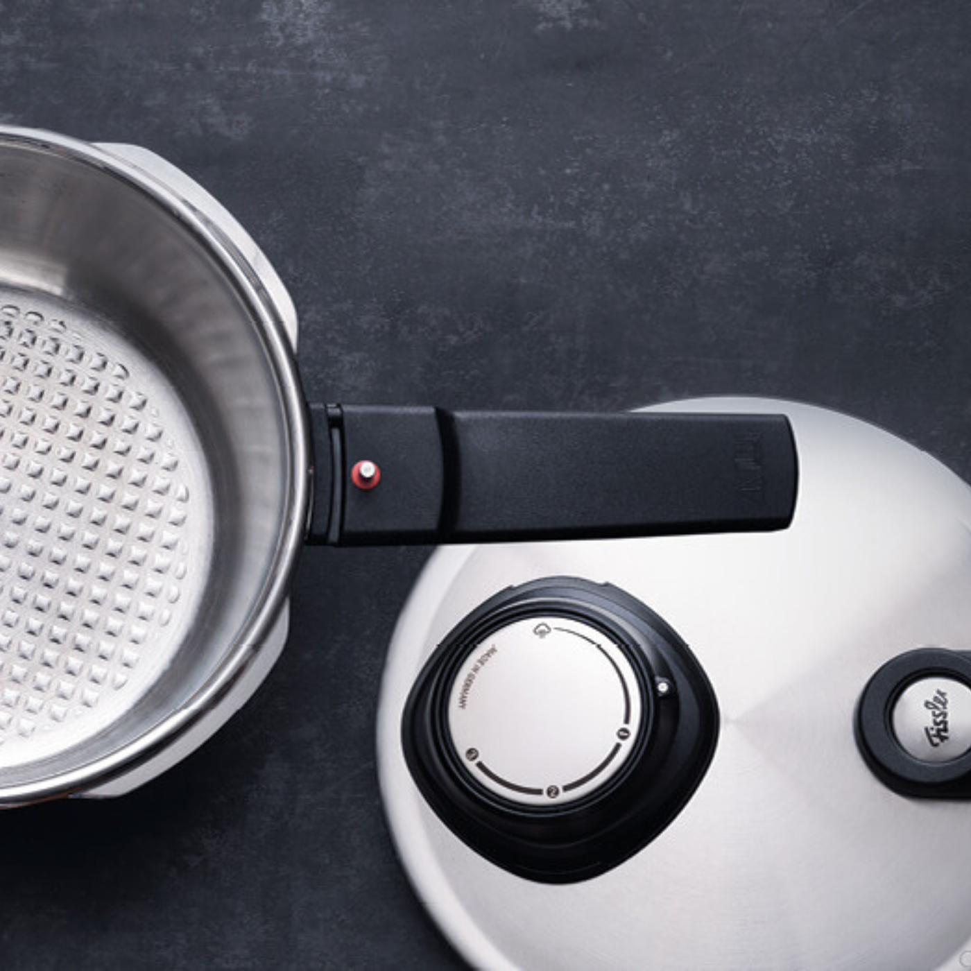 Fissler Schnellkochtopf aus rostfreiem Stahl mit einem charakteristischen strukturierten Boden, schwarzem Griff und Deckel auf einer dunklen Oberfläche.