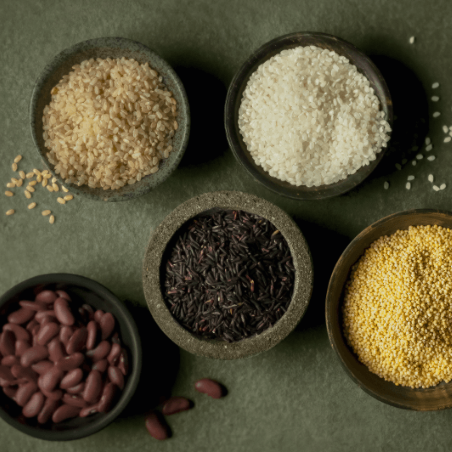 Fünf Schüsseln mit Körnern und Hülsenfrüchten - brauner, weißer und schwarzer Reis, Kidneybohnen, Hirse - sind in einem Fissler-Kochgeschirr auf Grün angeordnet.