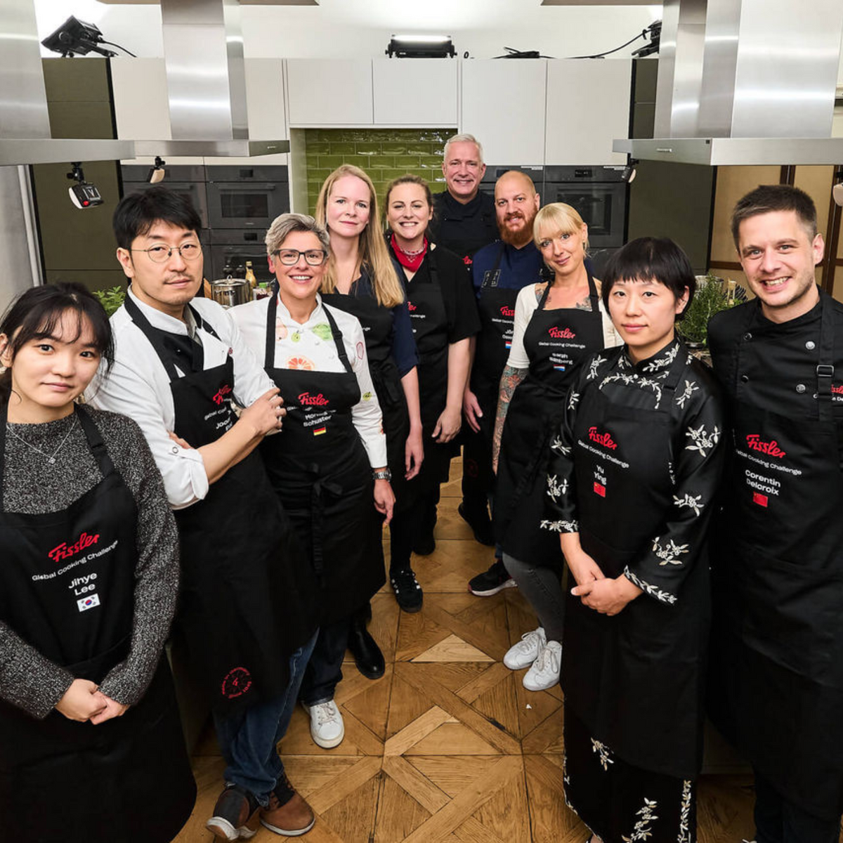 Ten people in black aprons smile together in an elegant kitchen featuring Fissler stainless steel cookware, exuding teamwork and innovation.