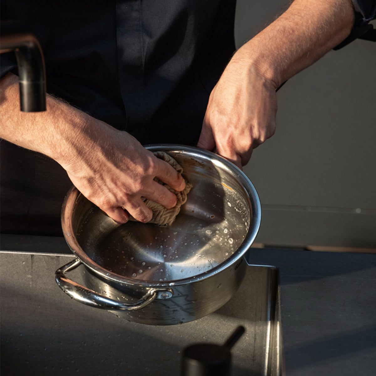 Eine Person reinigt die Fissler M5 Pro-Ply® Stielkasserolle mit Metalldeckel – in Deutschland aus recyceltem Edelstahl gefertigt – sorgfältig mit einem Schwamm am Spülbecken und spült Seife und Rückstände ab. Dieser auf Langlebigkeit und mühelose Pflege ausgelegte, spülmaschinenfeste Topf mit Induktionskompatibilität steht beispielhaft für Fisslers Engagement für Innovation, Nachhaltigkeit und zeitloses Design.
