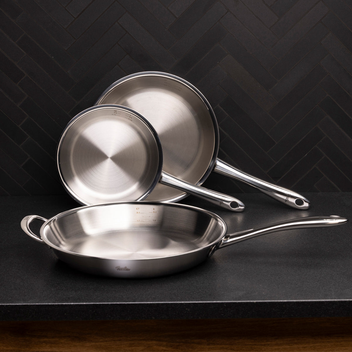 Drei Fissler M5 Pro-Ply®-Pfannen aus hochwertigem, recyceltem Edelstahl in verschiedenen Größen sind sorgfältig gestapelt und auf einer dunklen Arbeitsplatte vor einer eleganten, schwarzen Küchenrückwand mit Fischgrätenmuster angeordnet. Die zeitlose, langlebige Qualität und das präzise Design von Fissler – Made in Germany seit 1845 – stehen im Mittelpunkt dieses stilvollen Küchenensembles.