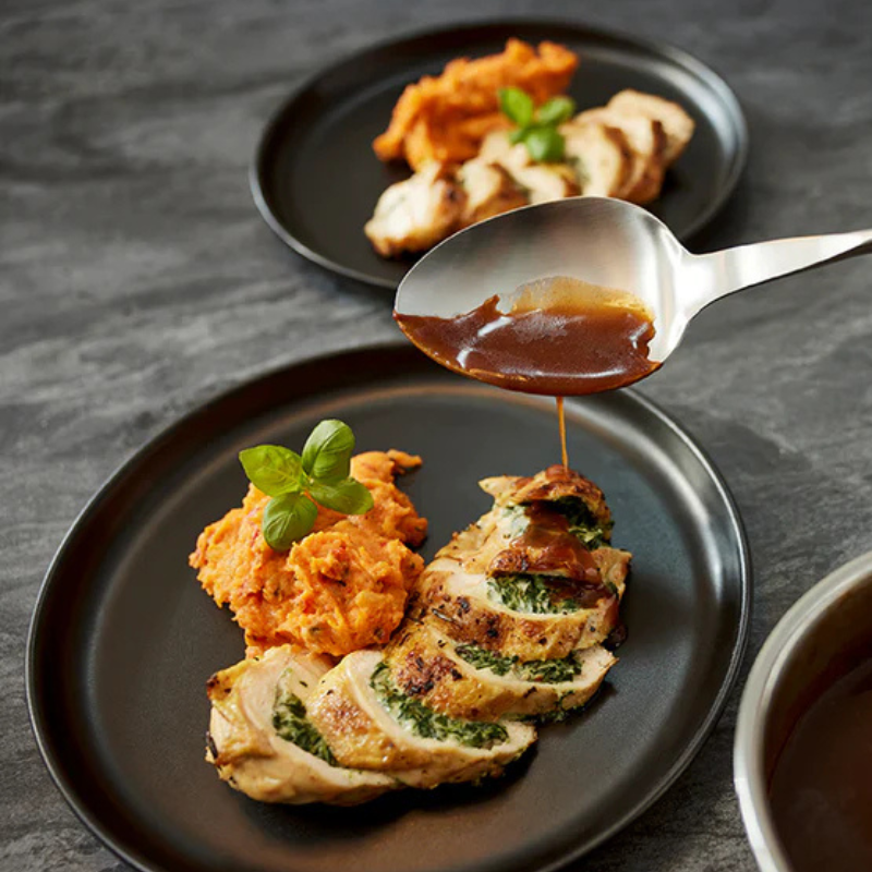 Eine Hand gießt Sauce über gefülltes Hähnchen und Süßkartoffelpüree, elegant angerichtet auf schwarzem Fissler-Kochgeschirr.