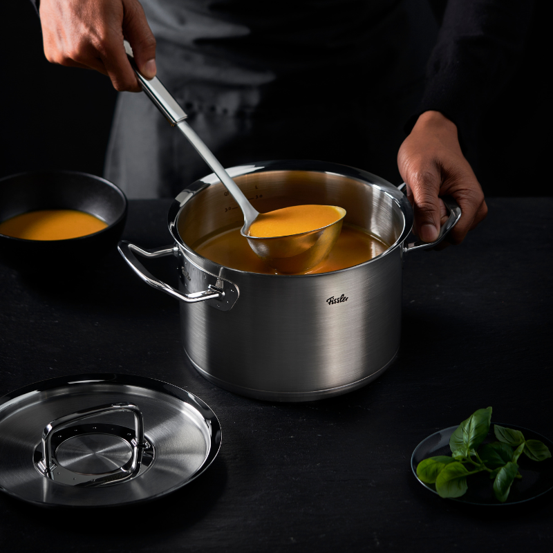 Eine Person schöpft Suppe aus einem hochwertigen Fissler-Edelstahltopf in eine Schüssel; Topfdeckel und Basilikum liegen auf einer dunklen, eleganten Arbeitsplatte.