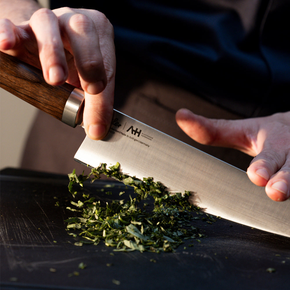 A person expertly chops fresh herbs on a sleek black cutting board using a premium, precision-engineered Fissler kitchen knife. Their fingers guide the blade with confidence and safety, reflecting Fissler’s heritage of innovation and timeless German quality in every detail.