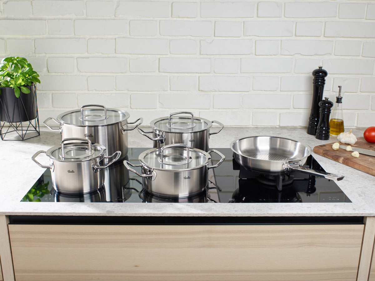Fissler Original-Profi Collection® 5-piece set on induction hob, white wall, cutting board with vegetables and spices.