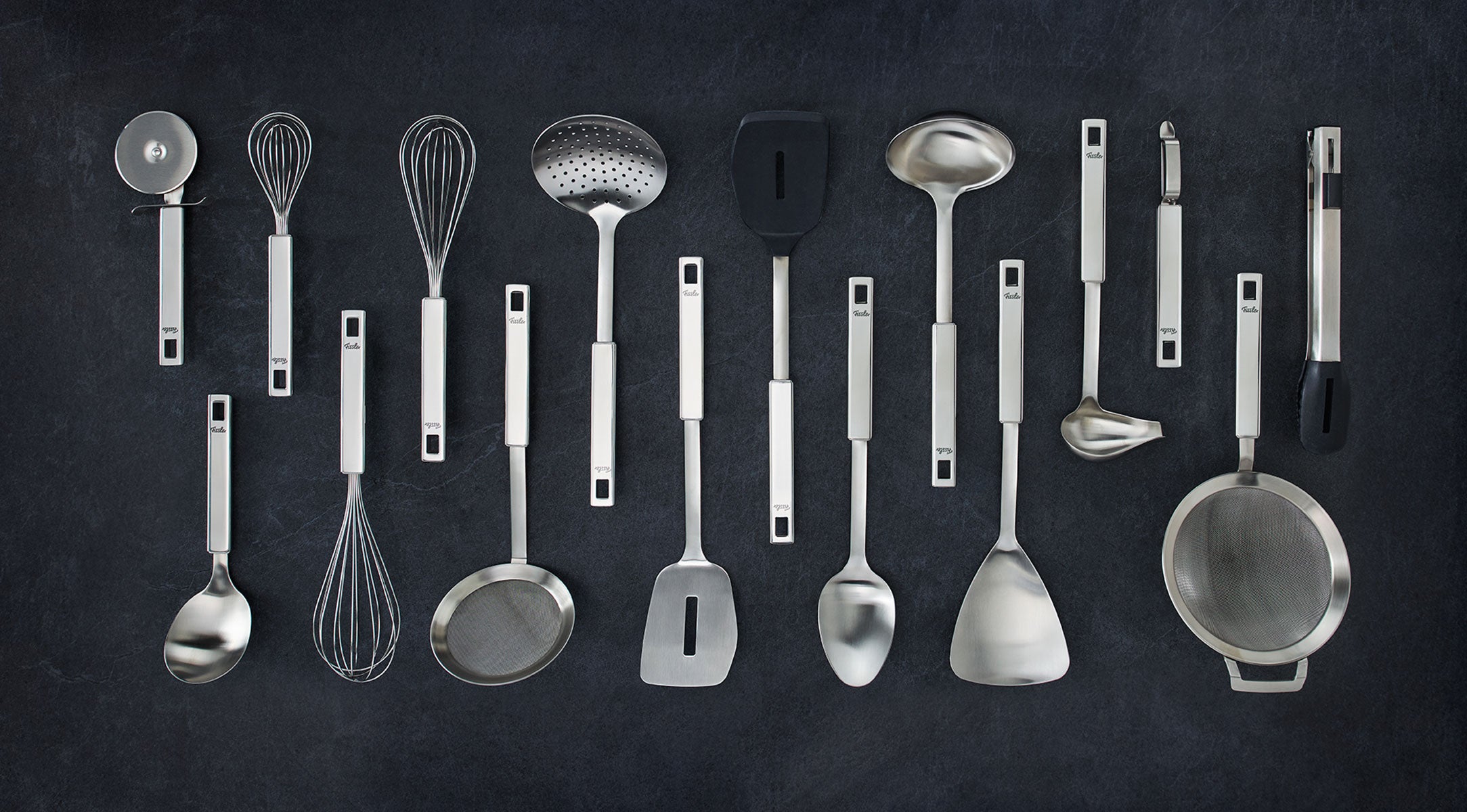 Küchenutensilien von Fissler aus rostfreiem Stahl - darunter Schöpfkellen, Pfannenwender und Schneebesen - sind auf einer dunklen Fläche in Reihen angeordnet.