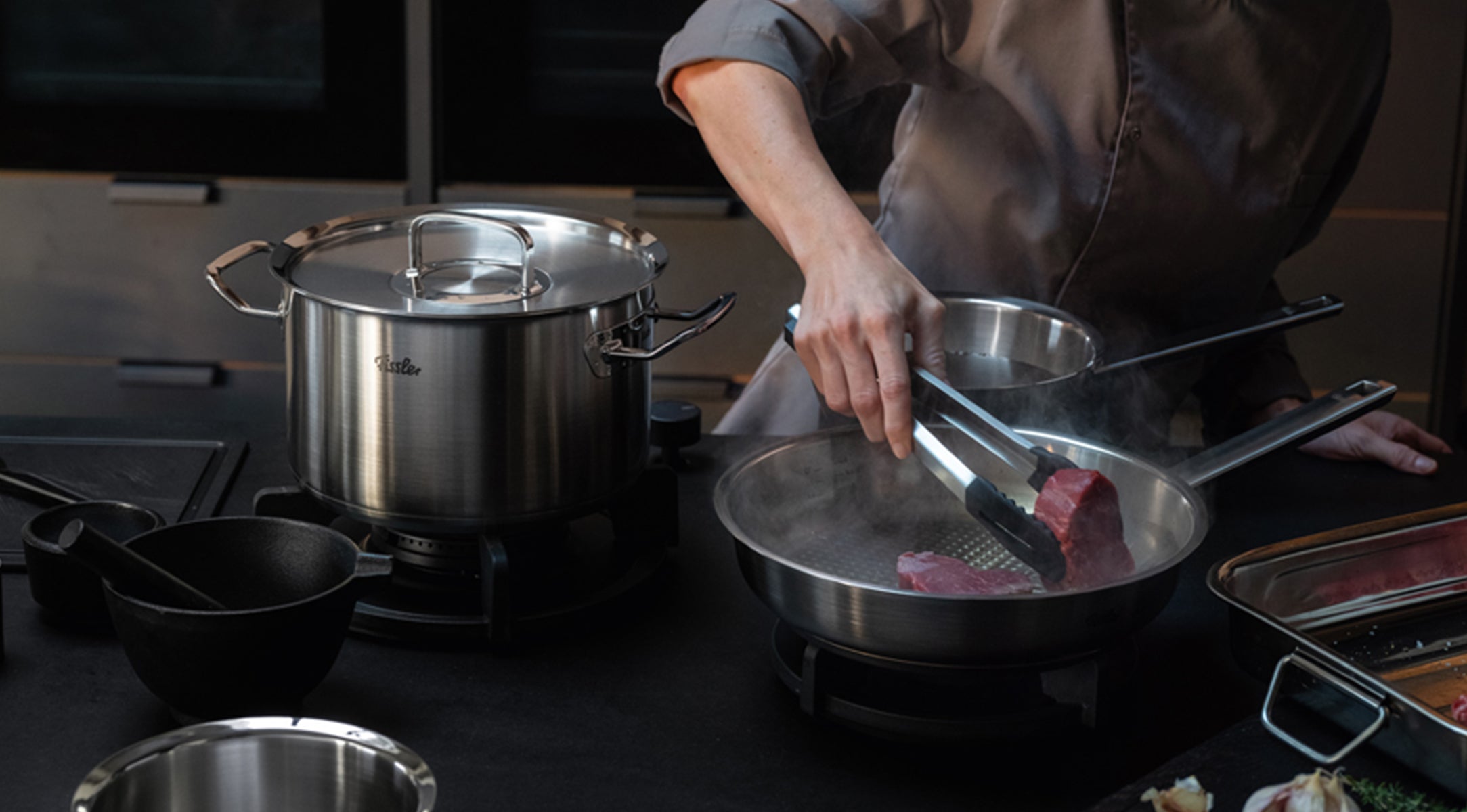 A chef in a professional jacket expertly sears a cut of meat in a Fissler stainless steel skillet using tongs. Beside them, a sizable Fissler pot sits on the stove, surrounded by premium kitchen utensils on an elegant dark countertop, showcasing the precision engineering and timeless design of Fissler cookware made in Germany.