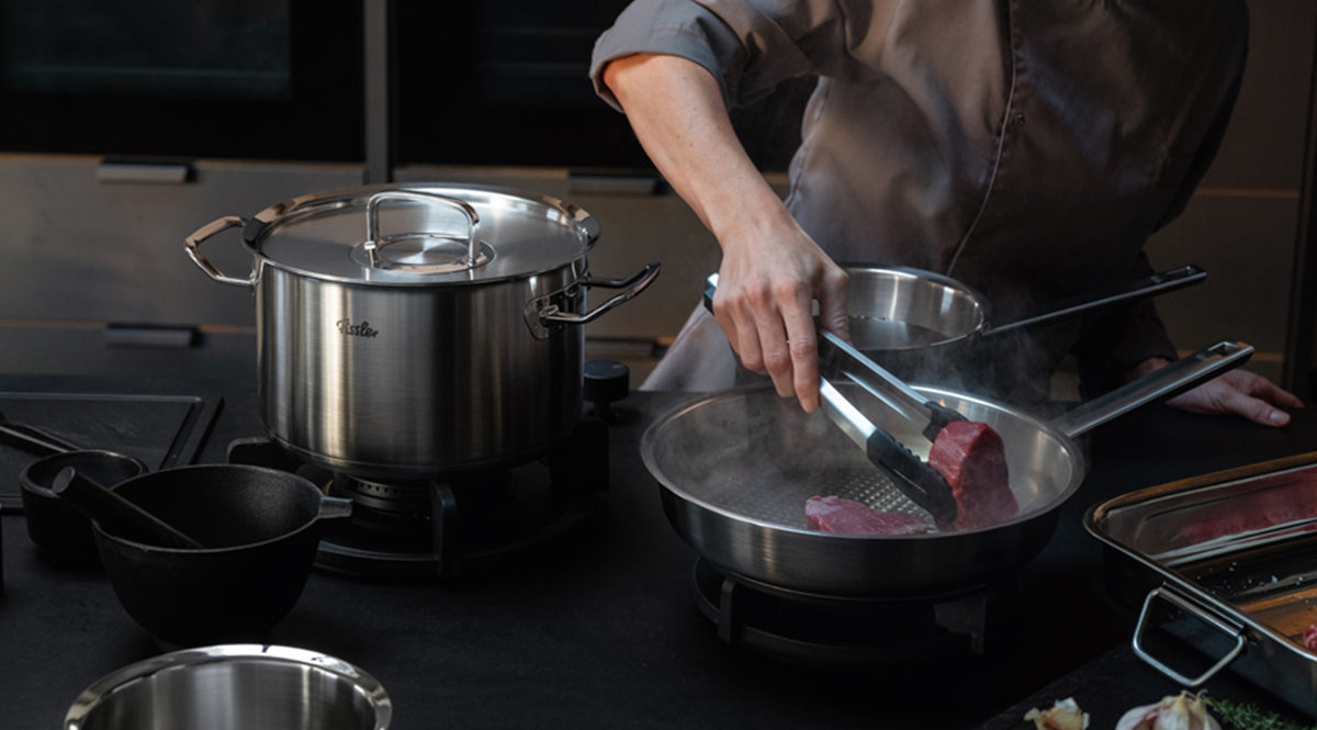 A chef in a white coat expertly sears a piece of meat using tongs in a premium Fissler stainless steel pan on the stove, while a matching Fissler pot and high-quality kitchen utensils, renowned for German precision and innovation, are arranged within reach.