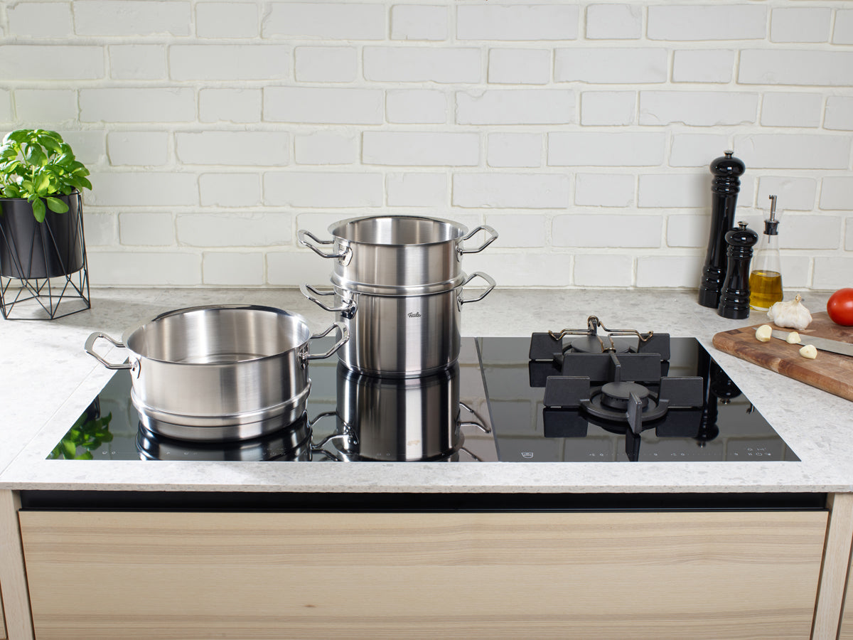 Zwei Fissler Original-Profi Collection® Töpfe und Dämpfeinsatz auf schwarzem Glaskochfeld; frische Zutaten unterstreichen deutsche Qualität.
