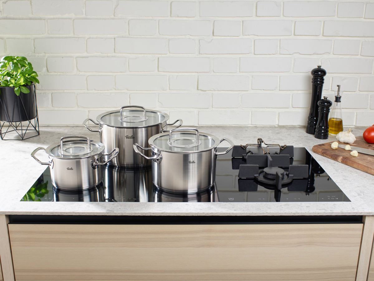 Drei Fissler Profi Collection® Edelstahltöpfe mit Glasdeckel auf Induktionsherd, daneben Gewürze, Gemüse und Zubehör.