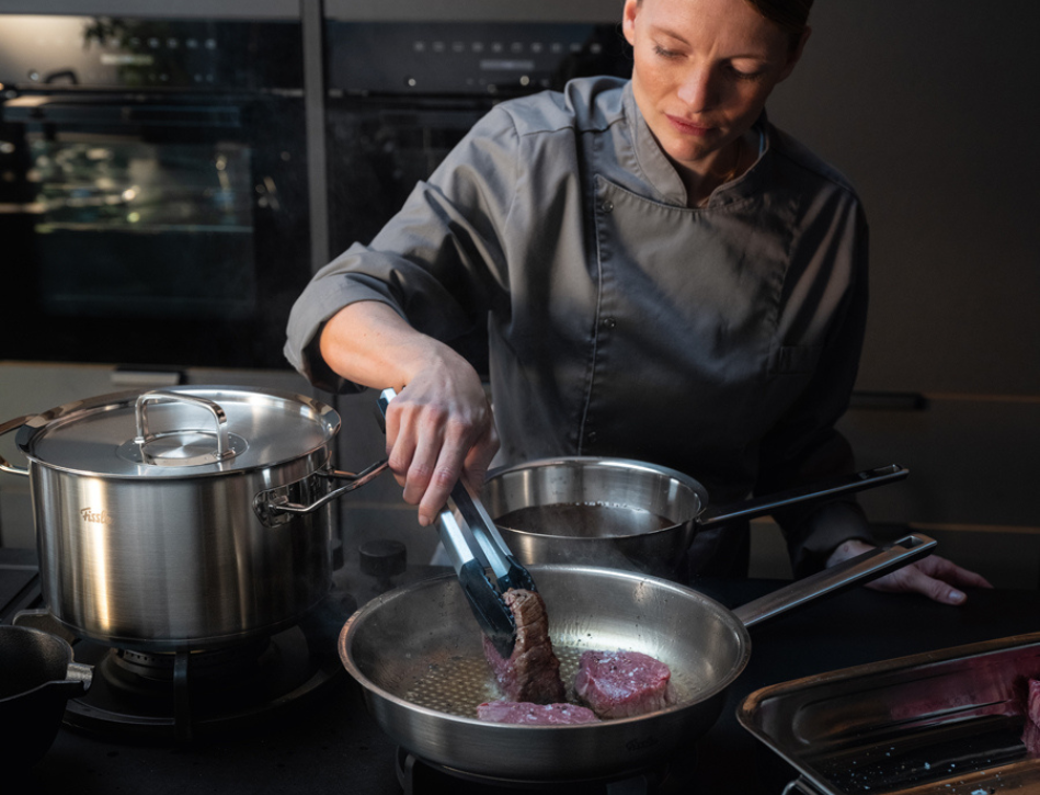 Ein Koch in Grau benutzt eine Zange zum Anbraten von Steaks in einer Fissler Bratpfanne mit Fissler Edelstahltopf und -pfanne in einer eleganten modernen Küche.