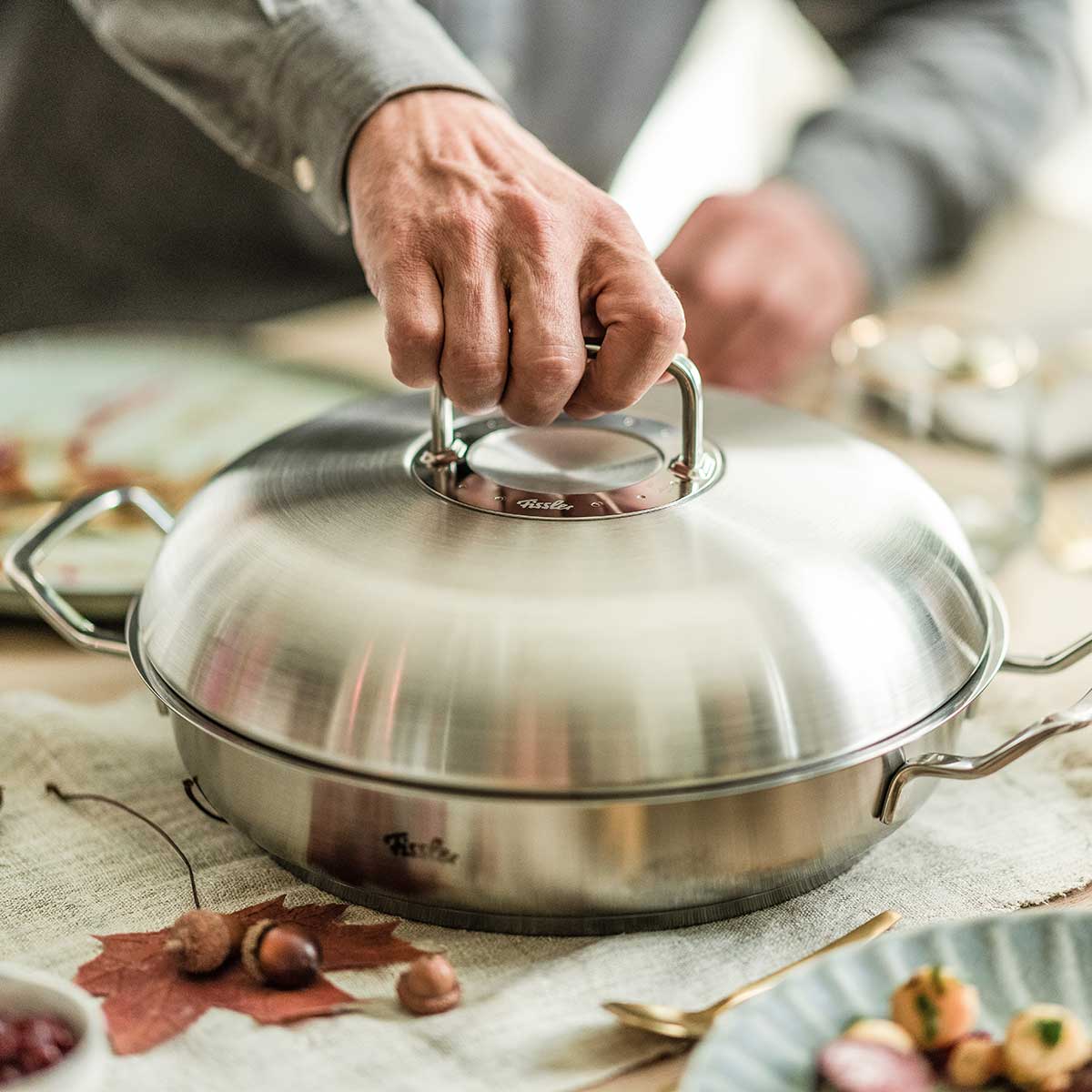 Auf einer festlichen Herbsttafel hebt eine Person den Deckel eines Fissler-Edelstahltopfes an, der deutsche Präzision und zeitloses Design zeigt.