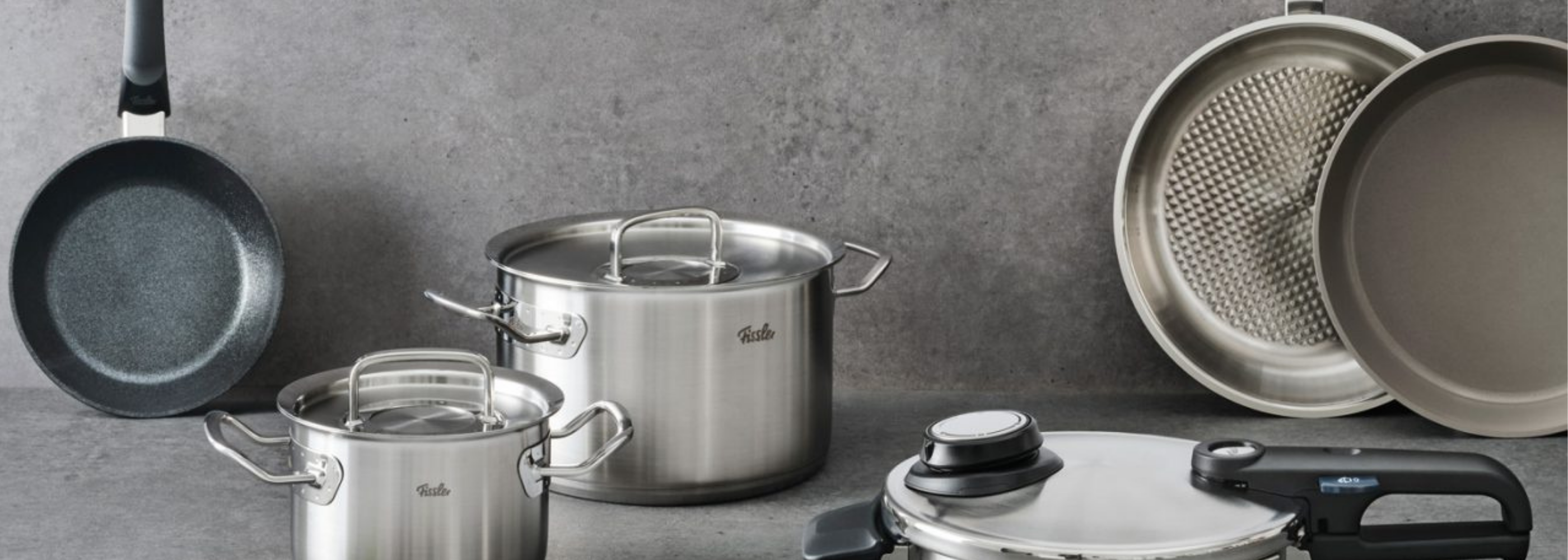 Fissler Edelstahltöpfe mit Deckeln, Bratpfanne, antihaftbeschichtete Bratpfanne und Schnellkochtopf auf grauer Oberfläche, zeitloses deutsches Design.