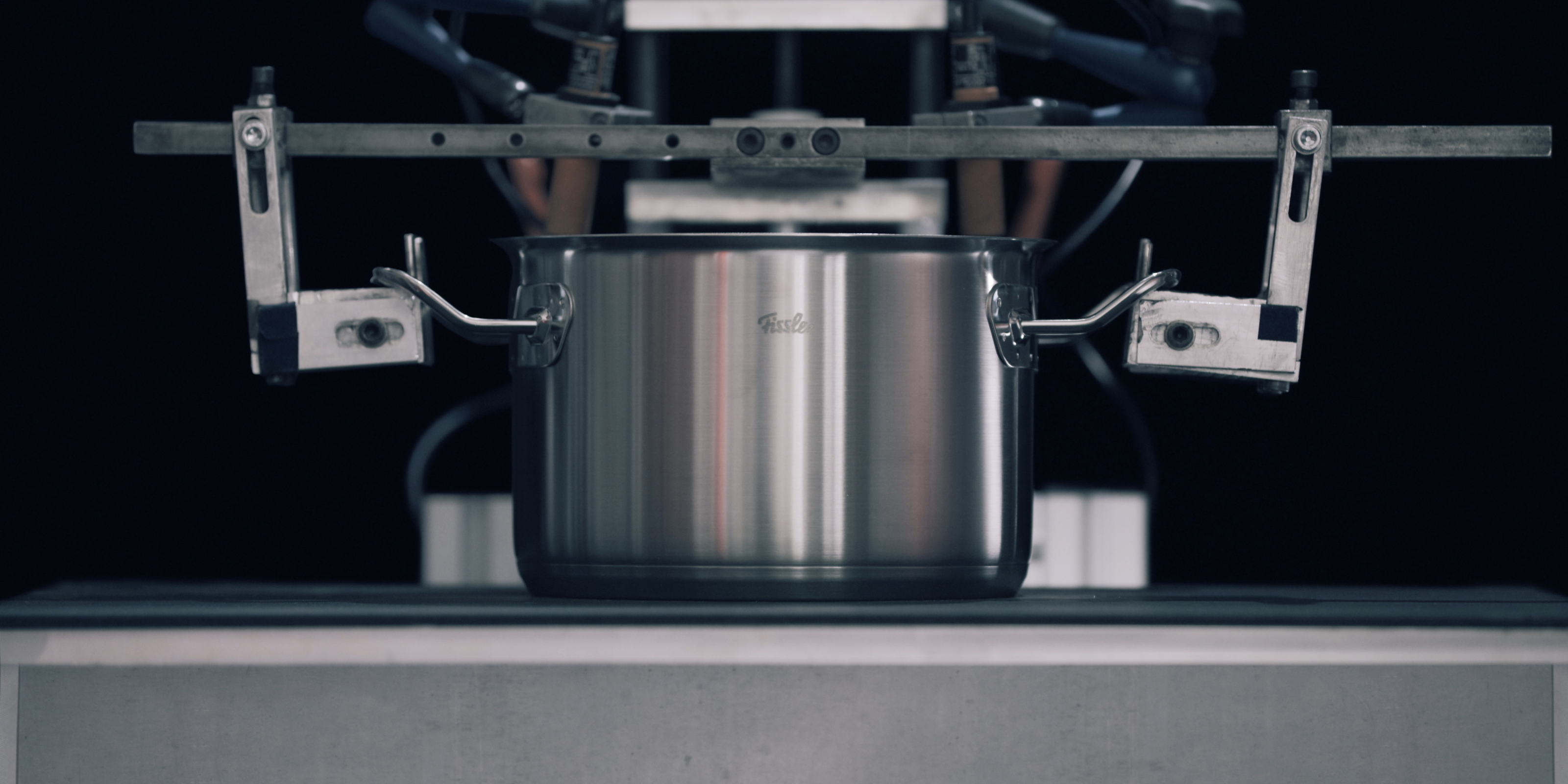 Ein Fissler-Edelstahltopf, der von zwei Klammern präzise gehalten wird, stellt die beständige deutsche Handwerkskunst vor einem eleganten dunklen Hintergrund zur Schau.