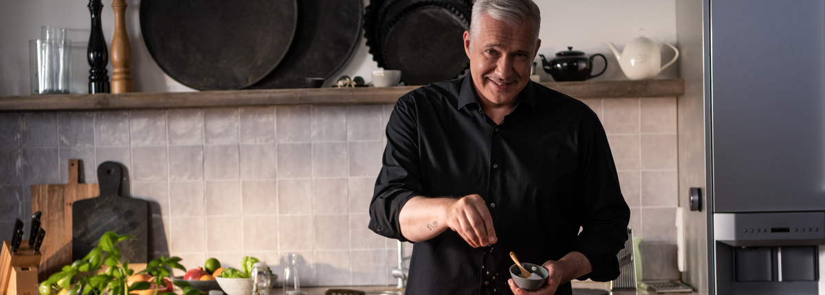 Ein Mann in einem schwarzen Hemd bereitet das Essen in einer modernen Küche mit hochwertigem Fissler-Kochgeschirr und frischem Gemüse auf der Theke zu.