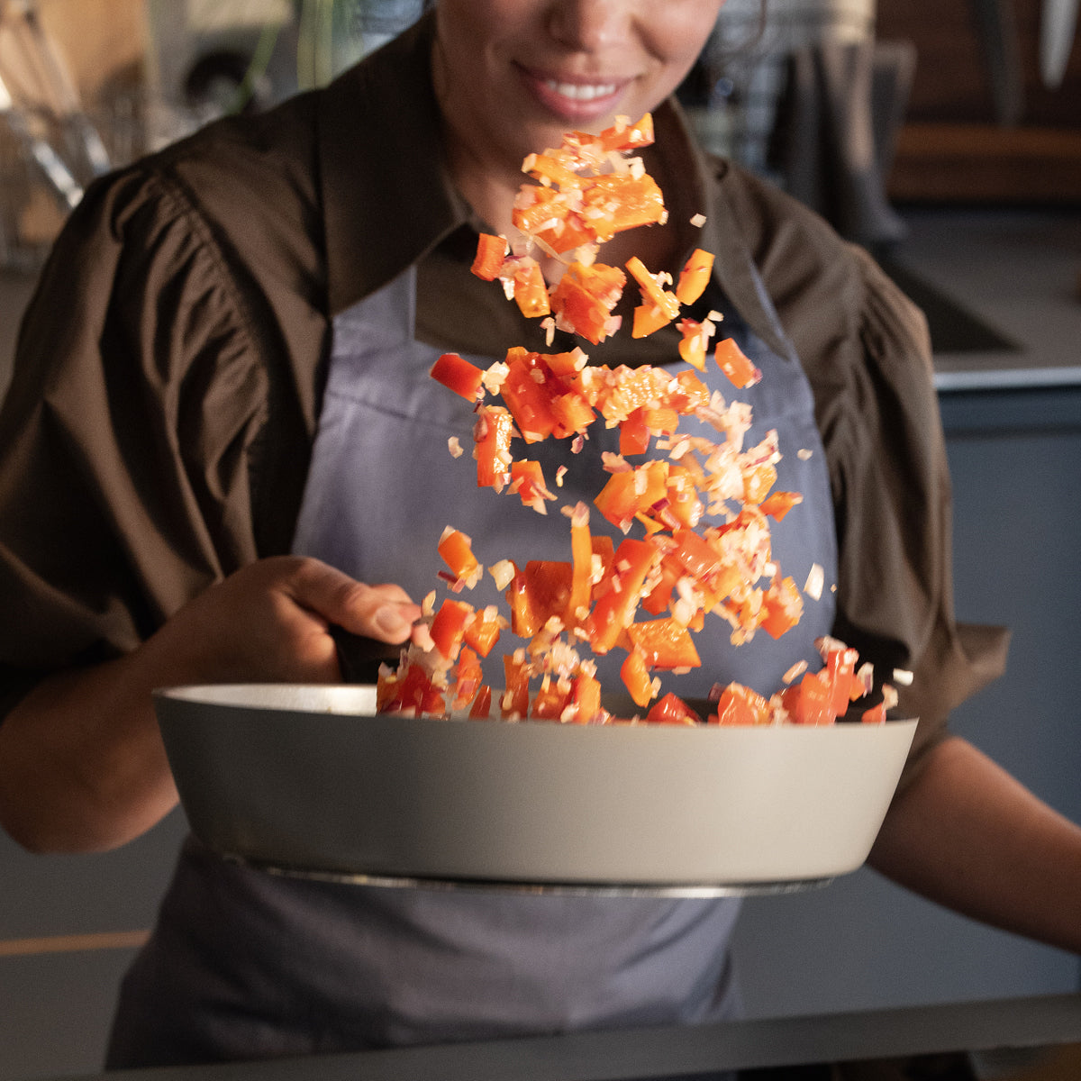 Ein Koch in einer Schürze schwenkt fachmännisch Paprika und Zwiebeln in einer Fissler-Edelstahlpfanne und stellt damit Präzision und Qualität unter Beweis.