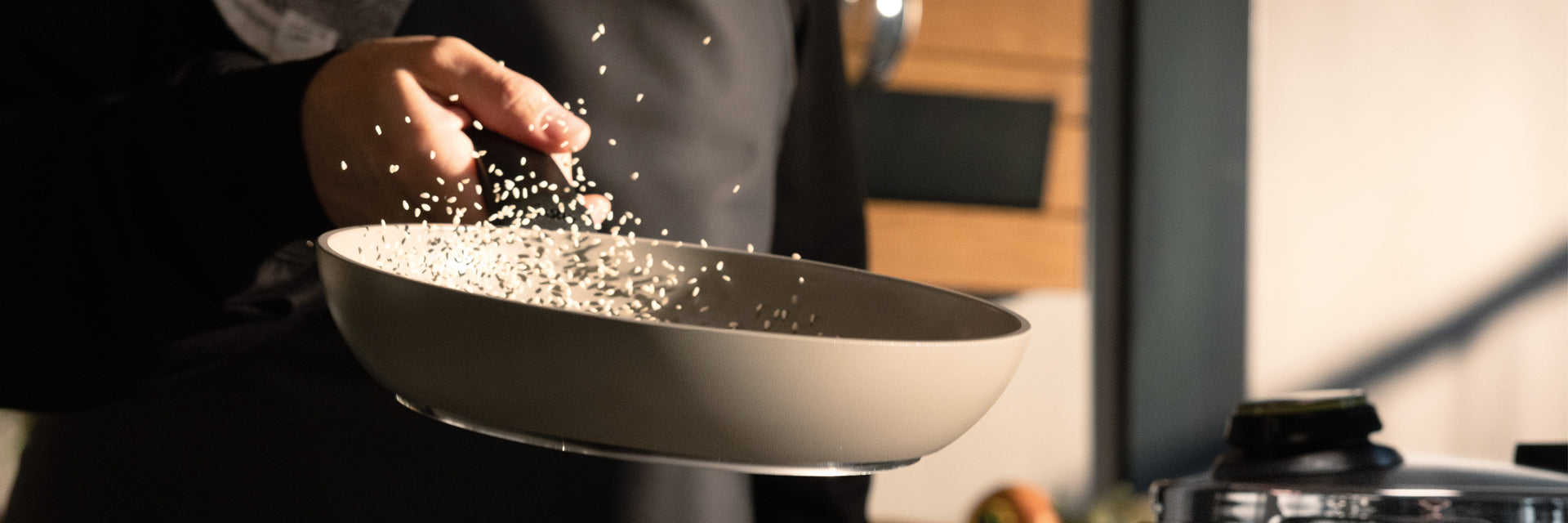 Ein Koch schwenkt gekonnt Reis in einer hochwertigen Fissler-Pfanne, wobei die Körner in der Luft tanzen. Die elegante Küchenszene verschwindet sanft im Hintergrund.