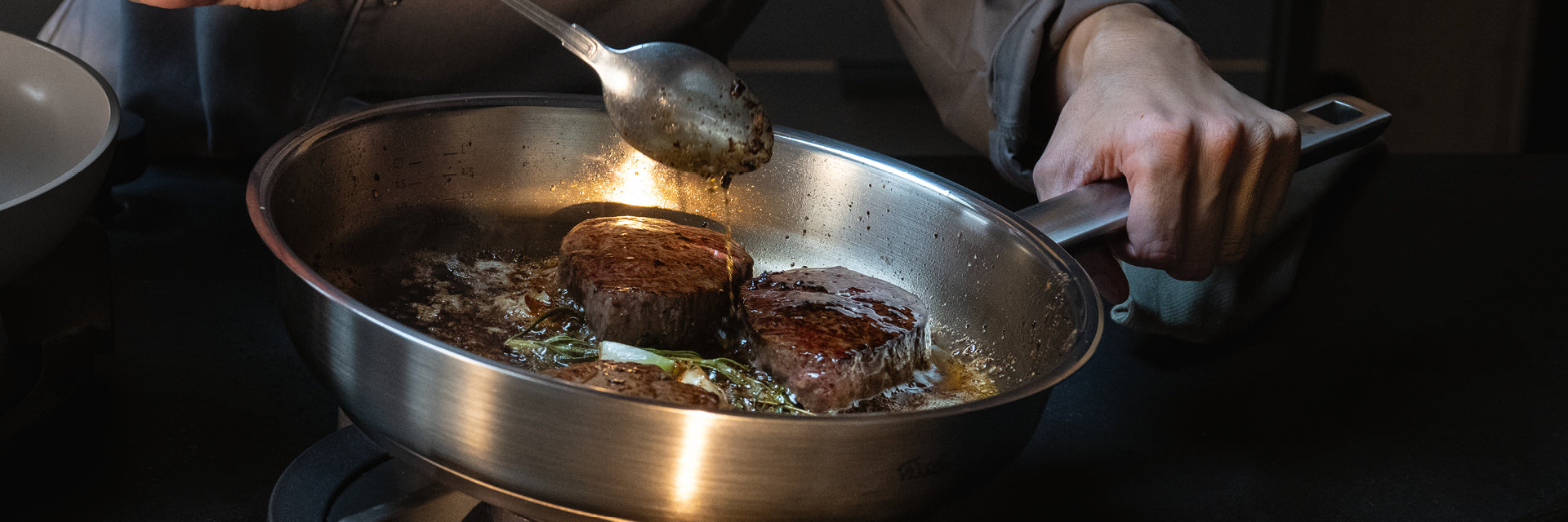 Eine Person brät zwei Steaks in einer Fissler-Edelstahlpfanne mit Kräutern und Butter und stellt damit unser deutsches Kochgeschirr auf dem Herd zur Schau.