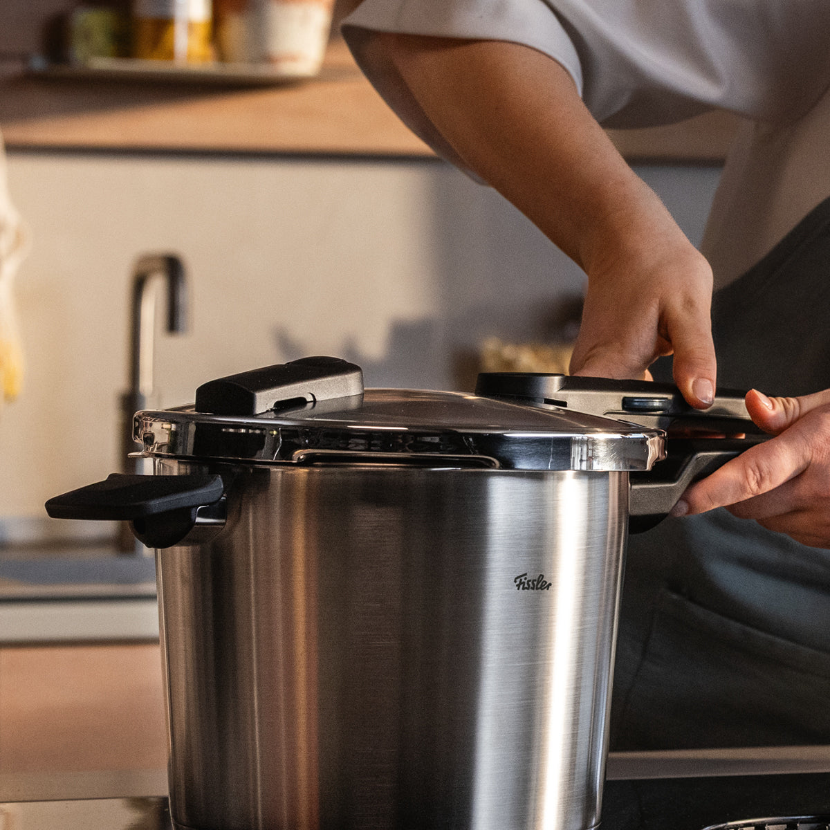 Eine Person in einer Küche schließt den Deckel eines Fissler Schnellkochtopfs aus Edelstahl, im Hintergrund sind Regale und ein Spülbecken zu sehen.