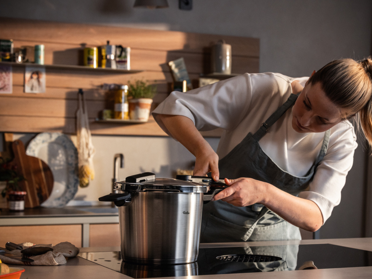A woman in an apron stands in a modern kitchen, attentively closing the lid of a Fissler stainless steel pressure cooker on the stove. Shelves displaying jars, plants, and kitchen accessories are seen in the background, highlighting the precision engineering and timeless design of Fissler cookware.
