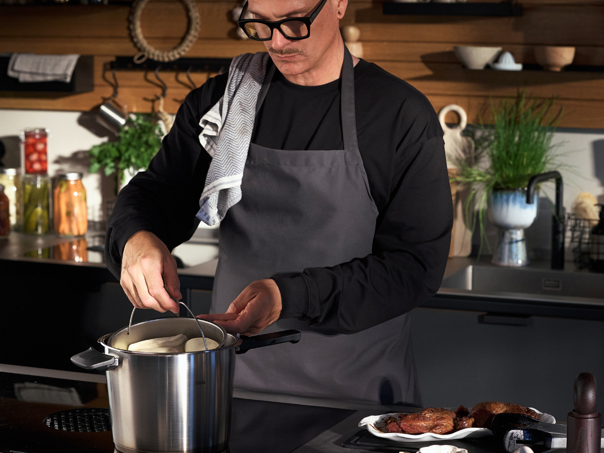 A person wearing glasses and a gray apron lifts the lid from a premium Fissler stainless steel pot on a stove in a sleek modern kitchen. On the counter, cooked meat rests elegantly on a plate beside the cookware, showcasing enduring quality and precision engineering.
