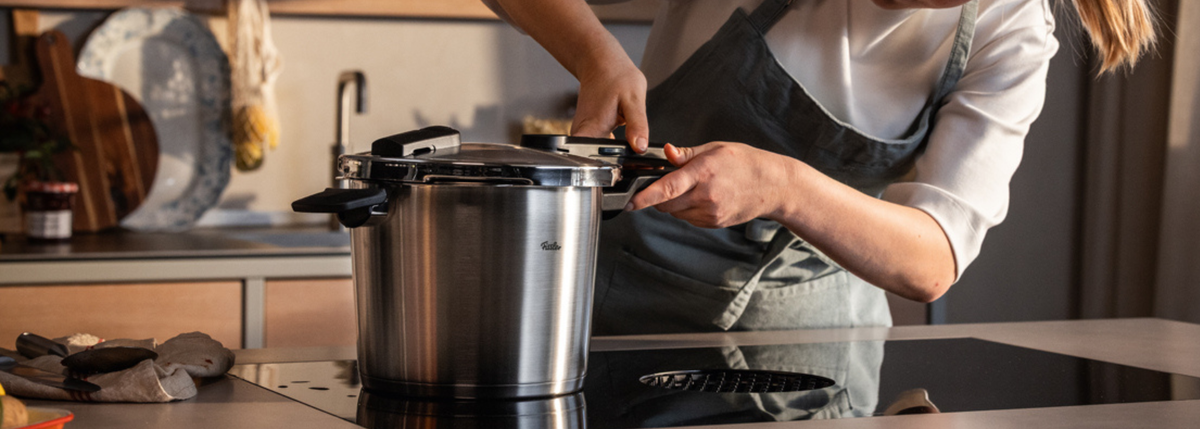 A person wearing an apron secures the lid on a Fissler stainless steel pressure cooker in a sleek, contemporary kitchen, highlighting the brand’s renowned German engineering and timeless design.