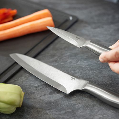 Fissler stainless steel knives on a gray countertop, one in hand. Sliced carrots and green vegetables on a black cutting board.