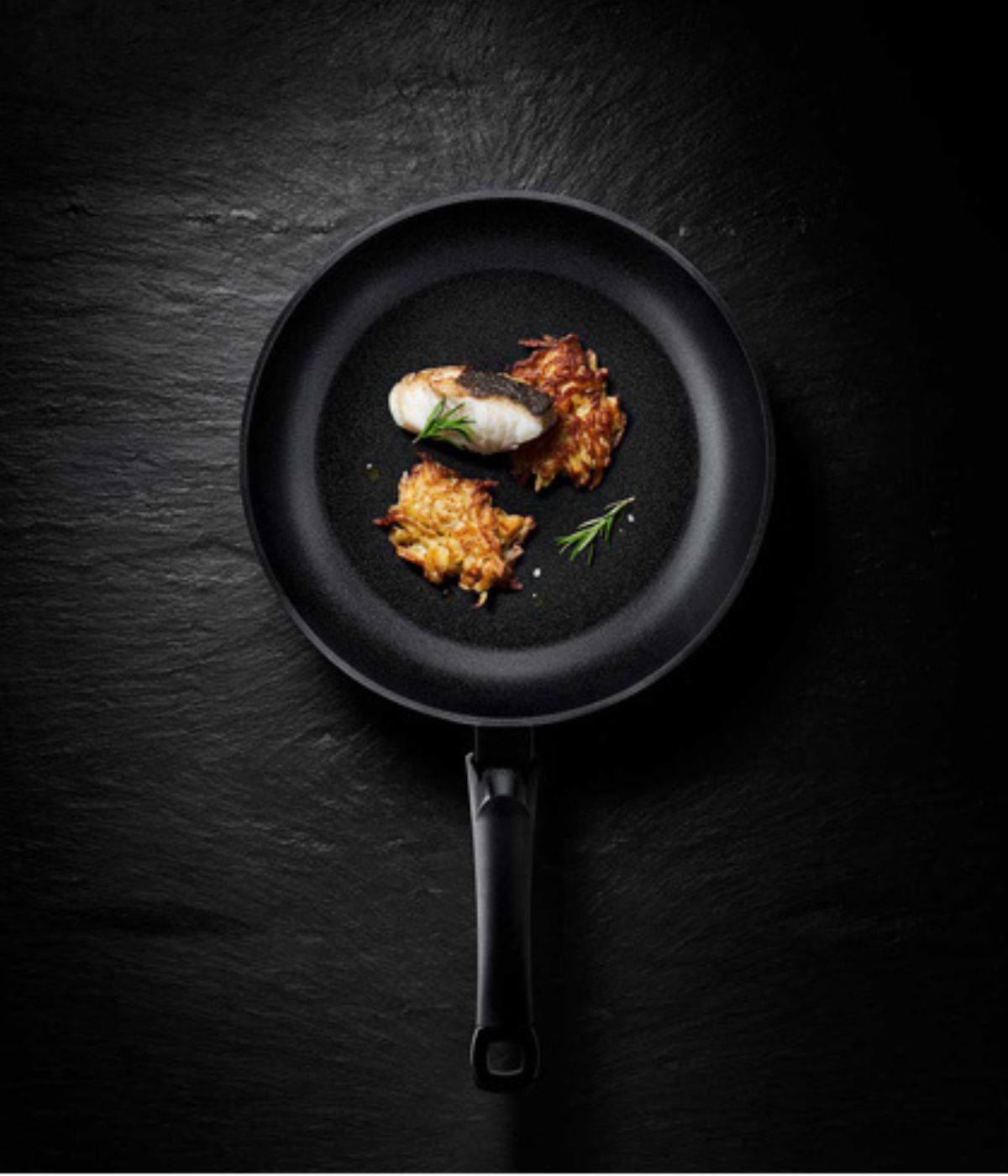Eine schwarze Bratpfanne von Fissler mit gekochtem Fisch, frischen Kräutern und zwei knusprigen Röstkartoffeln, die deutsche Wertarbeit widerspiegeln.