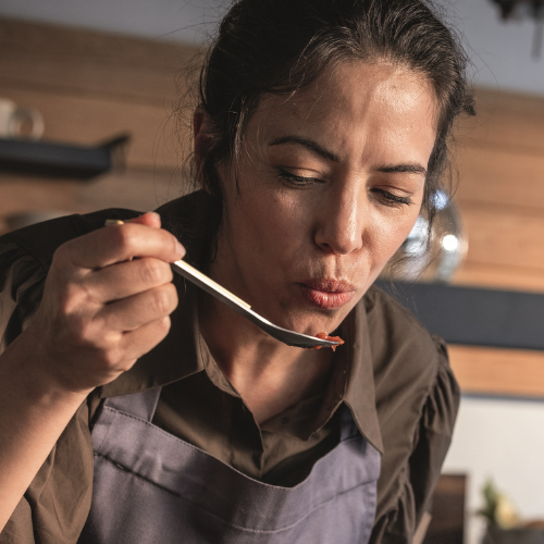 Eine Frau in einer grauen Schürze probiert mit einem Löffel, während sie in ihrer Küche mit hochwertigem Fissler-Edelstahlgeschirr kocht.