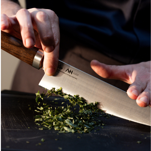 Eine Person hackt frische Kräuter auf einem schwarzen Schneidebrett mit einem Fissler-Küchenmesser mit einem präzisionsgefertigten Holzgriff fein.