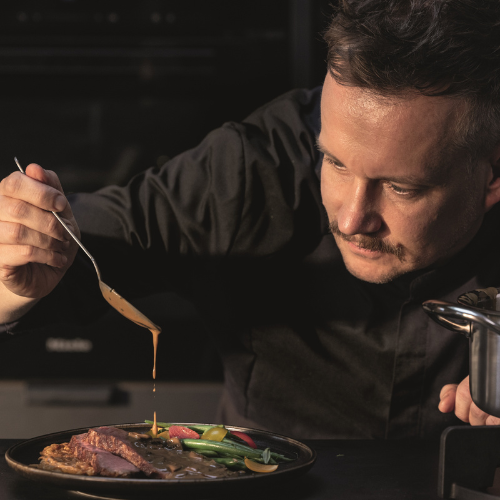 Ein Chefkoch bereitet ein Gourmetgericht mit Fissler-Kochgeschirr zu und beträufelt Fleisch und Gemüse in einer raffinierten, schummrigen Küche mit Sauce.