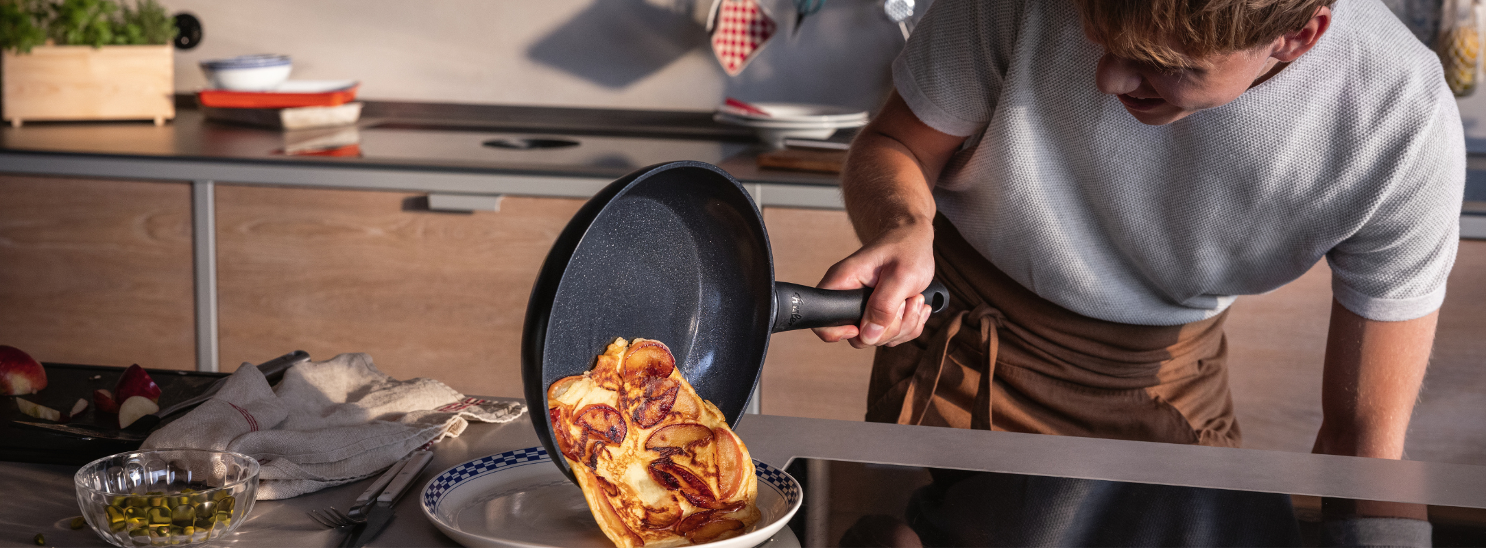 Eine Person gleitet fachmännisch ein goldenes Omelett aus einer Fissler-Pfanne auf einen Teller; hochwertiges Kochgeschirr und Utensilien stehen auf der Arbeitsplatte.