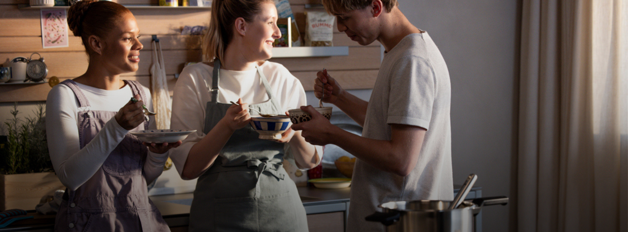 Drei Personen genießen eine Mahlzeit in einer sonnenbeschienenen Küche mit Fissler-Edelstahlkochgeschirr, das zeitlose Qualität und deutsches Design unterstreicht.