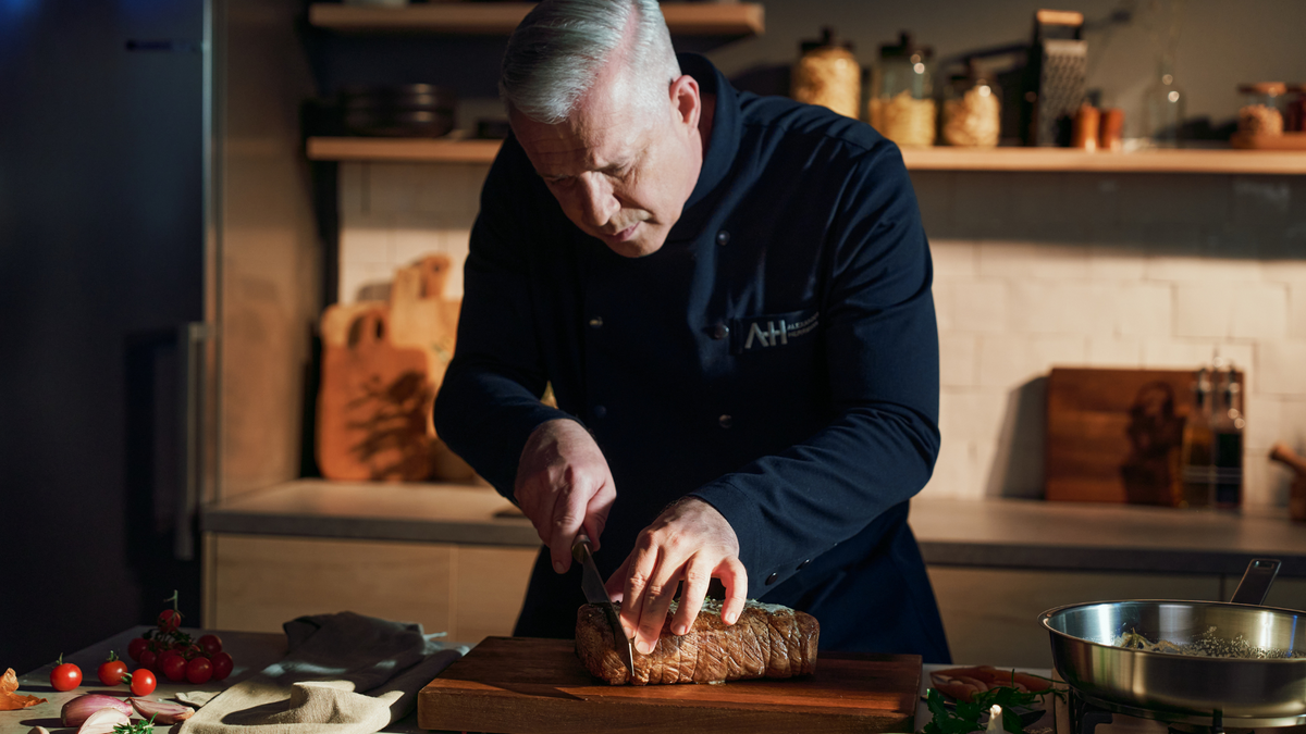 Ein Koch mit grauem Haar in dunkler Uniform schneidet in einer eleganten, modernen Küche mit Fissler-Kochgeschirr Braten auf einem Holzbrett.