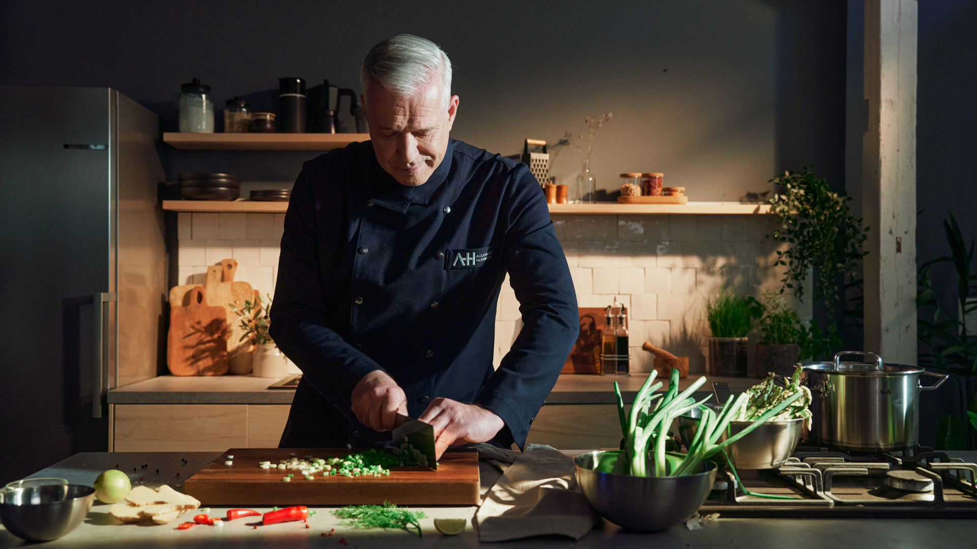 Ein Koch in dunkler Uniform schneidet grüne Zwiebeln auf einem Holzbrett, daneben steht rostfreies Kochgeschirr von Fissler und frisches Gemüse.