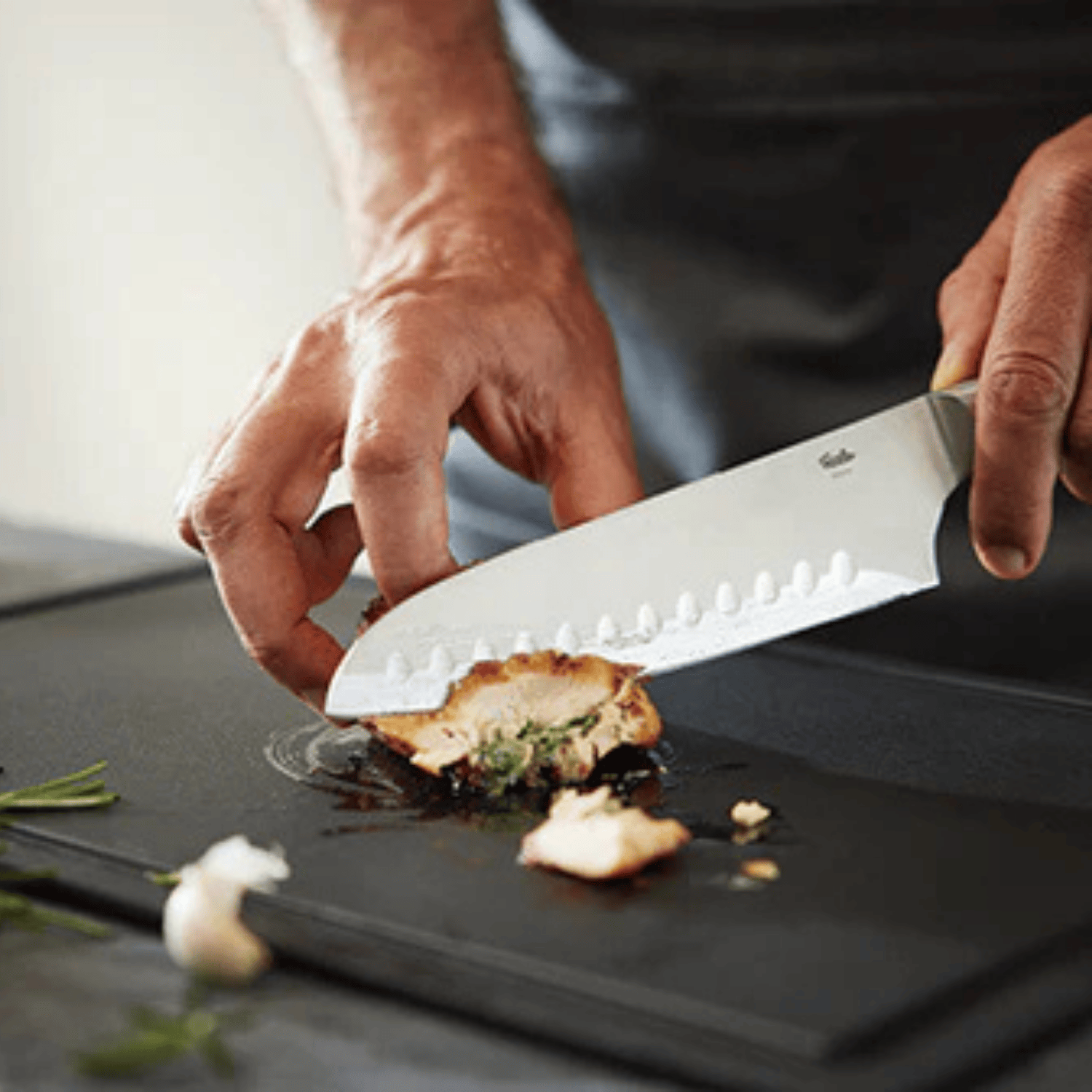 Eine Person schneidet gefüllte Hähnchenbrust mit einem präzisen Fissler-Kochmesser auf einem Schneidebrett auf, Kräuter und Knoblauch liegen daneben.